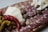 A charcuterie board featuring an assortment of sliced cured meats, including salami and prosciutto, alongside sliced cheese, pickled peppers, small meatballs, and onions. The arrangement creates a visually appealing display on a wooden board.