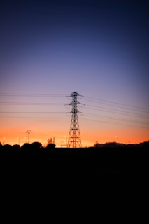 A silhouette of a large electrical transmission tower stands against the backdrop of a vibrant sunset. The sky transitions from a deep orange near the horizon to a dark blue higher up. Lights are visible in the distance, along with a few smaller utility poles and trees.