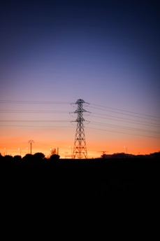 A silhouette of a large electrical transmission tower stands against the backdrop of a vibrant sunset. The sky transitions from a deep orange near the horizon to a dark blue higher up. Lights are visible in the distance, along with a few smaller utility poles and trees.