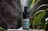 A small brown bottle with a dropper lid labeled 'CBD Kapljice' is situated on a light-colored wooden surface. The bottle features a label with green accents and appears to contain liquid. Surrounding the bottle are green leaves from a succulent plant, creating a natural setting. Wisps of white smoke or mist add an ethereal quality to the atmosphere.
