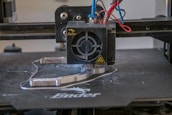 A 3D printer head actively printing on a flat surface, with visible heated printing element and extruding filament. Various colored wires are connected to the printing head.