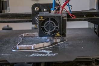 A 3D printer head actively printing on a flat surface, with visible heated printing element and extruding filament. Various colored wires are connected to the printing head.