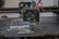 A 3D printer head actively printing on a flat surface, with visible heated printing element and extruding filament. Various colored wires are connected to the printing head.