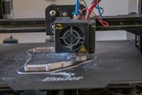 A 3D printer head actively printing on a flat surface, with visible heated printing element and extruding filament. Various colored wires are connected to the printing head.