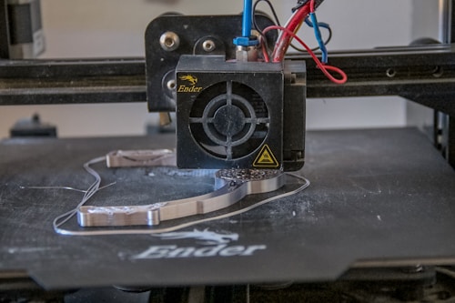 A 3D printer head actively printing on a flat surface, with visible heated printing element and extruding filament. Various colored wires are connected to the printing head.