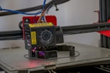 A close-up view of a 3D printer extruder in operation. The extruder has visible warning labels, multiple colored wires, and is positioned above a partially formed object being printed on the printer bed. The background includes some of the printer's frame and additional components.