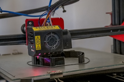 A close-up view of a 3D printer extruder in operation. The extruder has visible warning labels, multiple colored wires, and is positioned above a partially formed object being printed on the printer bed. The background includes some of the printer's frame and additional components.