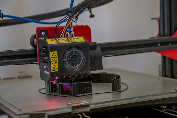A close-up view of a 3D printer extruder in operation. The extruder has visible warning labels, multiple colored wires, and is positioned above a partially formed object being printed on the printer bed. The background includes some of the printer's frame and additional components.