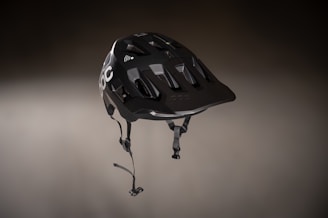 A sleek, black helmet with a modern design featuring angular vents and a matte finish. It is suspended in a slightly blurred, neutral background, highlighting its detailed construction. The helmet has a chin strap with clasps and logo branding visible on one side.