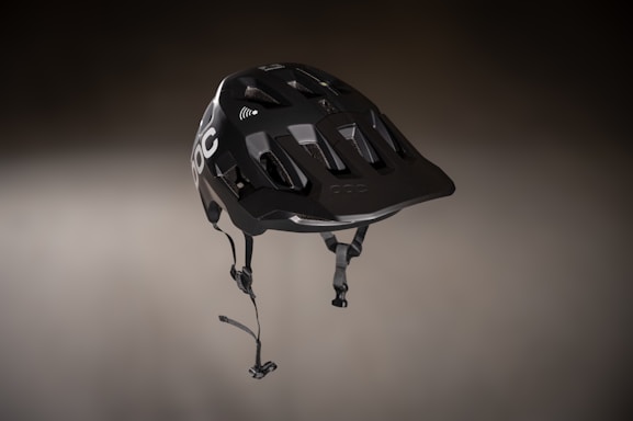 A sleek, black helmet with a modern design featuring angular vents and a matte finish. It is suspended in a slightly blurred, neutral background, highlighting its detailed construction. The helmet has a chin strap with clasps and logo branding visible on one side.