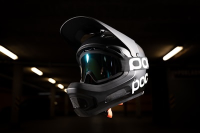 A black full-face helmet with integrated goggles, featuring the brand logo 'poc' on the side strap. The background is dark with minimal lighting, highlighting the helmet as the focal point.