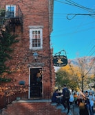 A brick building has ivy growing on its walls, and a sign reads 'The Pig Hill Inn Bed & Breakfast' hangs beside an entrance. Several people are gathered outside, some walking by while others are seated on the steps. The scene is set against a clear blue sky with autumn-colored trees.