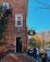 A brick building has ivy growing on its walls, and a sign reads 'The Pig Hill Inn Bed & Breakfast' hangs beside an entrance. Several people are gathered outside, some walking by while others are seated on the steps. The scene is set against a clear blue sky with autumn-colored trees.