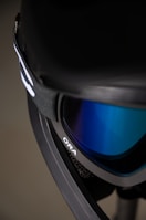 A close-up view of a ski or snowboard goggle with a black frame and blue reflective lenses. The strap is visible, as well as part of the goggle’s branding, which reads 'ORA'. The background is blurred, bringing focus to the goggles.