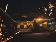 A traditional Japanese-style house illuminated at night with warm lights, creating a cozy atmosphere. Sparkles or fireworks highlight the foreground, adding a festive touch to the serene environment.