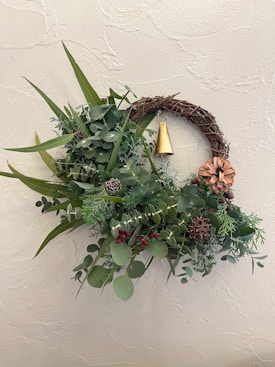 A decorative wreath made of green foliage, brown twigs, and small pine cones, adorned with a golden bell and a dried flower. Red berries are scattered throughout the greenery, creating a natural and festive appearance. The wreath is mounted on a textured light beige wall.
