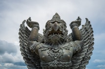 A large, intricately detailed statue of a mythological creature with an eagle-like face, wings, and muscular human body. The creature has its arms raised, and its expression is fierce, set against a cloudy sky.