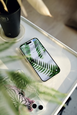 A smartphone with a clear case displaying green fern leaves on its screen is placed on a white surface. Next to it, there is another phone case with a similar botanical design featuring green and pink patterns. A dark plant pot is partially visible in the upper corner, suggesting a natural theme and lighting.