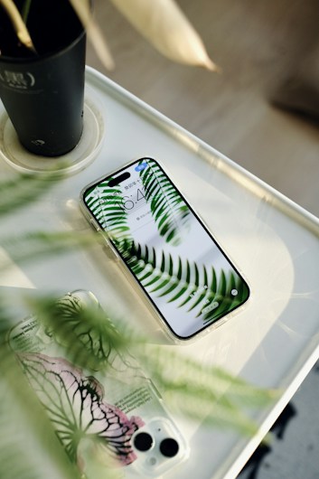 A smartphone with a clear case displaying green fern leaves on its screen is placed on a white surface. Next to it, there is another phone case with a similar botanical design featuring green and pink patterns. A dark plant pot is partially visible in the upper corner, suggesting a natural theme and lighting.