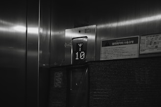 A dimly lit elevator interior with a floor indicator displaying the number 10. The surroundings include a metallic surface and a few signs with Korean writing on them.