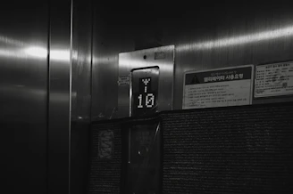 A dimly lit elevator interior with a floor indicator displaying the number 10. The surroundings include a metallic surface and a few signs with Korean writing on them.