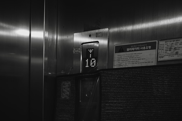 A dimly lit elevator interior with a floor indicator displaying the number 10. The surroundings include a metallic surface and a few signs with Korean writing on them.