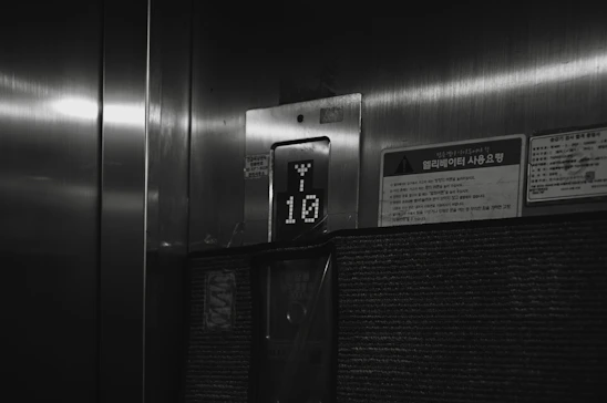 A dimly lit elevator interior with a floor indicator displaying the number 10. The surroundings include a metallic surface and a few signs with Korean writing on them.