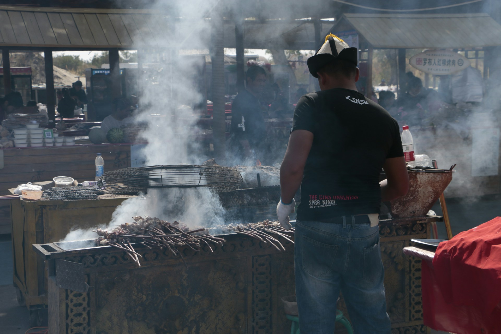 A man is standing at a smoky outdoor grill with skewers lined up, cooking food. He is wearing casual clothes and a hat. In the background, there are other stalls and people, suggesting a market or fair atmosphere. Various cooking tools and a bottle are visible nearby, contributing to a busy scene.