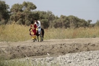 Two people are riding a horse along a dirt path surrounded by lush green vegetation and trees. One person is dressed in a vibrant red and yellow traditional outfit, while the other wears casual clothing. The setting appears rural and natural, with a peaceful and open atmosphere.