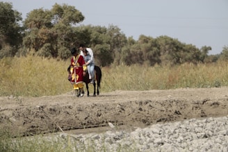 Two people are riding a horse along a dirt path surrounded by lush green vegetation and trees. One person is dressed in a vibrant red and yellow traditional outfit, while the other wears casual clothing. The setting appears rural and natural, with a peaceful and open atmosphere.