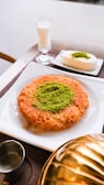 A plate features a traditional dessert made of shredded pastry with a center garnish of ground pistachios. Next to it is a glass of milk and a small square white dessert also topped with pistachios. The setting includes a metal container and a small jug, possibly for syrup.