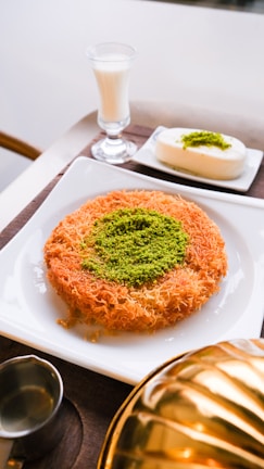 A plate features a traditional dessert made of shredded pastry with a center garnish of ground pistachios. Next to it is a glass of milk and a small square white dessert also topped with pistachios. The setting includes a metal container and a small jug, possibly for syrup.
