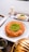 A plate features a traditional dessert made of shredded pastry with a center garnish of ground pistachios. Next to it is a glass of milk and a small square white dessert also topped with pistachios. The setting includes a metal container and a small jug, possibly for syrup.