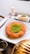 A plate features a traditional dessert made of shredded pastry with a center garnish of ground pistachios. Next to it is a glass of milk and a small square white dessert also topped with pistachios. The setting includes a metal container and a small jug, possibly for syrup.