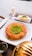 A plate features a traditional dessert made of shredded pastry with a center garnish of ground pistachios. Next to it is a glass of milk and a small square white dessert also topped with pistachios. The setting includes a metal container and a small jug, possibly for syrup.