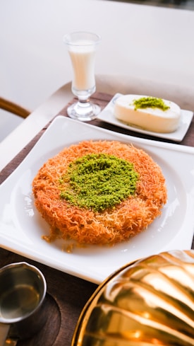 A plate features a traditional dessert made of shredded pastry with a center garnish of ground pistachios. Next to it is a glass of milk and a small square white dessert also topped with pistachios. The setting includes a metal container and a small jug, possibly for syrup.
