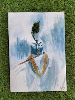 A watercolor painting depicts a serene, abstract figure with a peacock feather adorning the head. The figure has closed eyes and a calming presence, with a flute resting on the shoulder and a garland draped around the neck. Soft blues dominate the background, creating a soothing atmosphere.