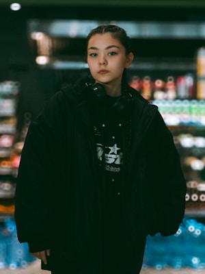 A person stands in a store aisle wearing a black jacket and headphones around their neck. The background features shelves stocked with a variety of products, including colorful bottles and packages.