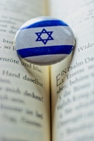 A Star of David pin rests on an open book with printed text in German. The pin features blue and white colors and contrasts against the beige pages, which show a depth of field with a sharp focus on the pin and blurring to the edges.