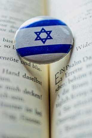 A Star of David pin rests on an open book with printed text in German. The pin features blue and white colors and contrasts against the beige pages, which show a depth of field with a sharp focus on the pin and blurring to the edges.