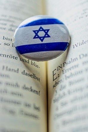 A Star of David pin rests on an open book with printed text in German. The pin features blue and white colors and contrasts against the beige pages, which show a depth of field with a sharp focus on the pin and blurring to the edges.
