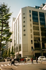 A bustling urban scene features a large office building with signage in an Asian script. People cross the street at a zebra crossing, with several vehicles waiting, including a van and a car. Trees line the sidewalk, and the sky is clear, creating a bright and lively atmosphere.
