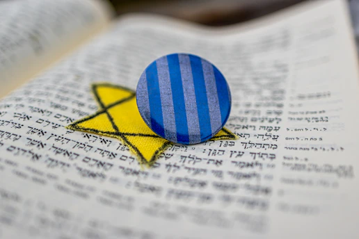 A blue and gray striped button rests atop a yellow Star of David on an open book with Hebrew text. The arrangement is sharp and in focus against a blurred background, highlighting the button and star.