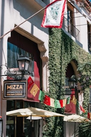 A charming street view featuring a building adorned with green ivy climbing the walls. There's a sign for an Italian restaurant named 'Mizza' with festive red, yellow, and green banners decorating the facade. Above, a flag flutters near a balcony. Outdoor umbrellas create a welcoming shaded area beneath.