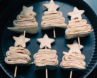 Six pastry pieces shaped like stylized Christmas trees are arranged in rows on a dark baking tray. Each piece comprises layered pastry folded in a zig-zag pattern, topped with a star and held together by a skewer.