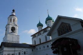 The image depicts a traditional Eastern Orthodox church with distinctive architectural features. It has multiple domed towers with green tops, each capped with a cross. The front facade of the building includes detailed religious iconography and artwork. The structure is primarily white with some dark roofing and intricate window designs. The sky is clear with a few clouds, indicating it is a sunny day.