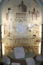The image features an exhibition display showcasing architectural drawings and photographs of Eastern Orthodox churches with distinctive domes and intricate architectural details. In the foreground, white stone artifacts are placed on a glass shelf, suggesting historical or archaeological significance.