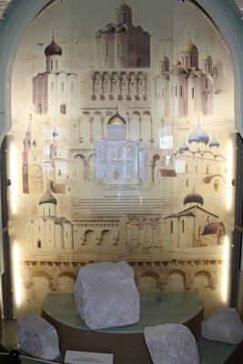 The image features an exhibition display showcasing architectural drawings and photographs of Eastern Orthodox churches with distinctive domes and intricate architectural details. In the foreground, white stone artifacts are placed on a glass shelf, suggesting historical or archaeological significance.