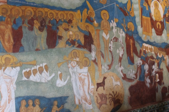 A detailed fresco depicting religious figures with halos, including angels blowing trumpets and various scenes of people in robes. There are animals, scales, and saintly figures looking solemn. The artwork has rich colors and intricate details, capturing a sense of divine judgment or ascension.