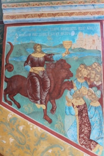 A historical painting depicts a figure holding a chalice, riding a multi-headed beast. Several people dressed in elaborate garments are bowing in the foreground. The background includes a body of water and a distant cityscape.